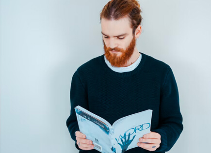 Man with a red beard in a dark sweater reading a magazine against a light blue wall.