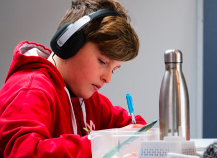 Student wearing headphones and a red hoodie writes with a blue pen at a desk, with a water bottle nearby in a study setting.